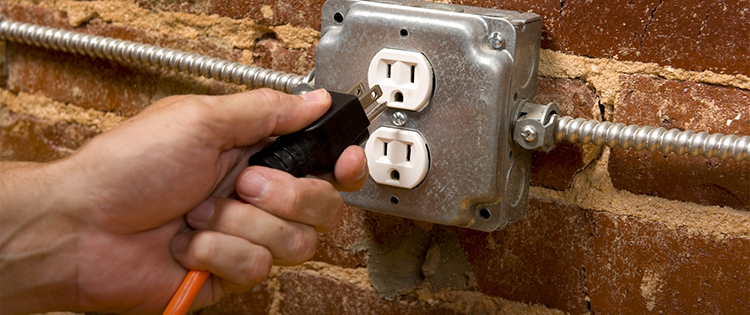 A hand plugging a power cord into an electrical outlet mounted on a brick wall